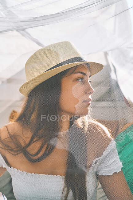 Though transparent curtain of content young female in sunhat chilling in backyard tent on sunny day and looking away — Stock Photo