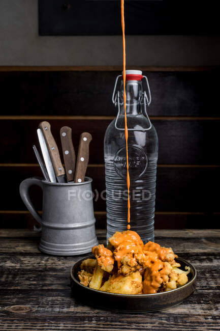 Сырный соус выливается на вкусную хрустящую курицу, помещенную на тарелку возле стеклянной бутылки с водой в темном ресторане — стоковое фото