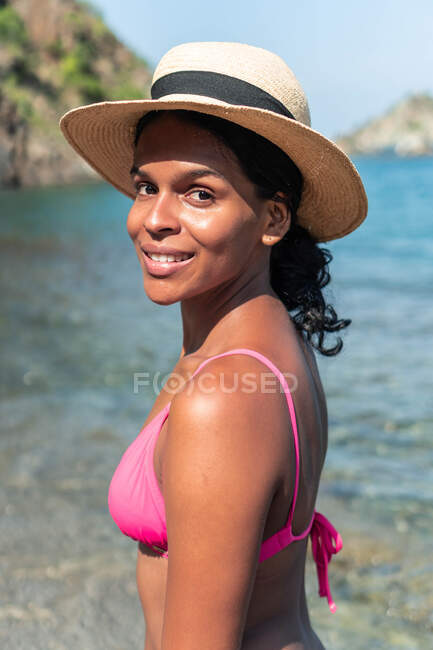 Side view of cheerful ethnic female tourist in swimwear and hat looking at camera on ocean coast — Stock Photo