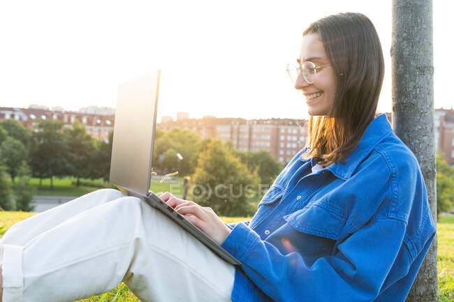 Side view of happy female freelancer leaning on tree and browsing netbook while working on project remotely in urban park on sunny day — Stock Photo