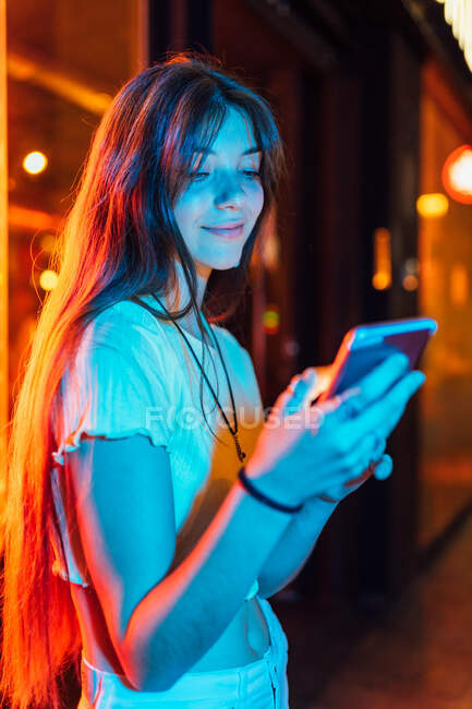 Цікава молода жінка в сережках, що серфінгують Інтернет на мобільному телефоні проти блискучих неонових ламп в місті — стокове фото
