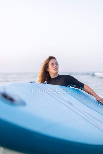Женщина в купальнике стоит с доской SUP в морской воде летом и смотрит в сторону — стоковое фото