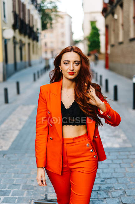 Решительная женщина в стильном ярко-оранжевом костюме трогает длинные рыжие волосы и гуляет по улицам города, глядя в камеру — стоковое фото