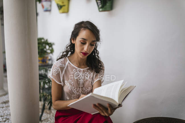 Content ethnic female in stylish outfit sitting on stool in patio and reading novel in book while enjoying summer weekend — Stock Photo