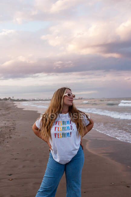 Positive young female in trendy sunglasses and stylish outfit standing at seaside against sea in summer evening — Stock Photo