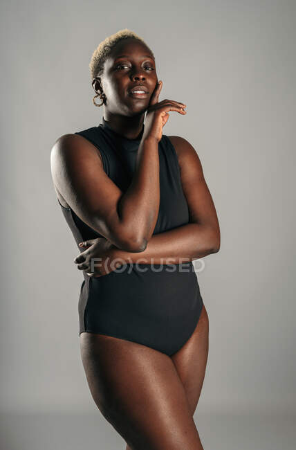 African American curvy female in black bodysuit standing with her hand over her face on gray background in studio — Stock Photo