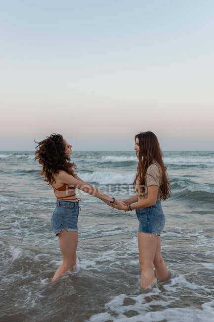 Seitenansicht junger Frauen, die Händchen halten, während sie beim romantischen Date in Meereswellen vor wolkenlosem Abendhimmel stehen — Stockfoto