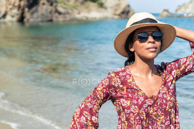 Ethnic female tourist in sundress standing with hand on head on sandy coast against ocean and mounts in sunlight — Stock Photo
