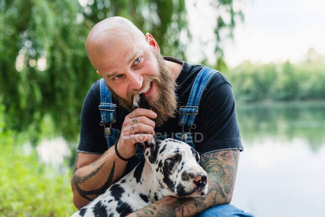 Bald male with beard in casual outfit sitting and playing with obedient spotted Great Dane puppy on lake with green trees on summer day — Stock Photo