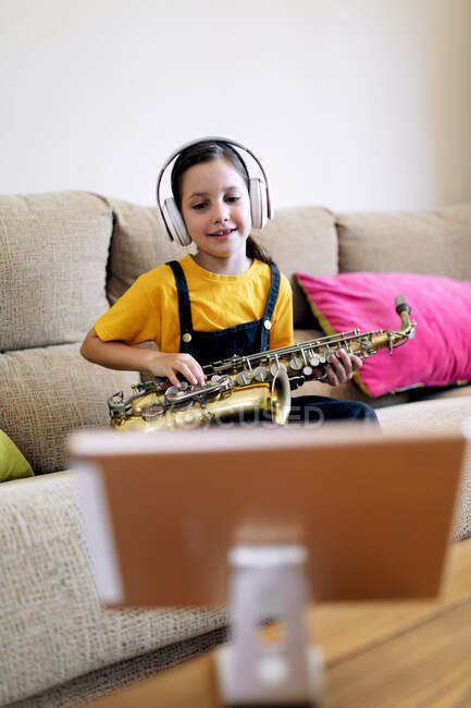 Enfant attentif dans un casque et saxophone sur le canapé enregistrement vidéo sur téléphone portable à la maison — Photo de stock