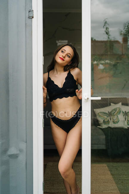 Teneur femelle mince en lingerie noire debout près de la porte en verre menant au balcon et regardant loin — Photo de stock