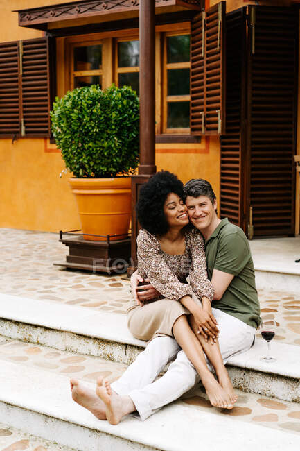 Positiva coppia multirazziale seduta su scale di pietra di portico di cottage e abbracciando mentre godendo il fine settimana insieme — Foto stock