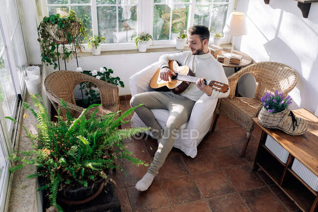 Musicista maschile contemplativo con tatuaggi che suonano la chitarra classica seduto in poltrona e guardando lontano contro la finestra in casa — Foto stock