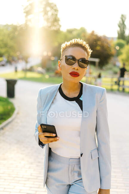 Portrait de la femme afro-américaine élégante en lunettes de soleil en utilisant un téléphone portable tout en se promenant sur la passerelle dans le parc urbain en regardant la caméra — Photo de stock