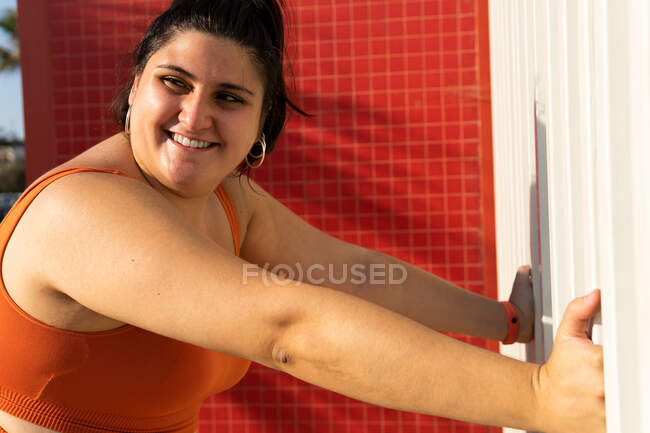 Sonriendo más tamaño atleta femenina étnica haciendo ejercicio mientras se apoya en la pared y mirando hacia otro lado en la calle soleada - foto de stock