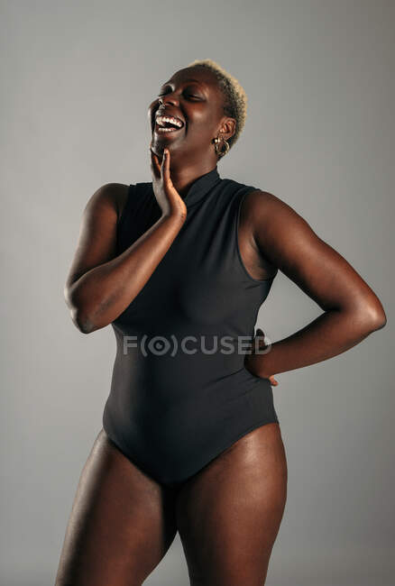 Cheerful African American curvy female in black bodysuit standing on gray background in studio and laughing — Stock Photo