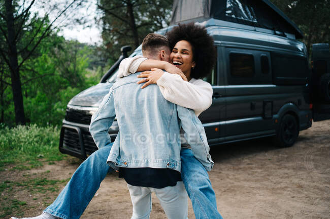Fröhliche junge Afroamerikanerin lacht glücklich und umarmt Freund, während sie während der gemeinsamen Sommerreise in der Nähe eines im grünen Wald geparkten Wohnmobils Spaß hat — Stockfoto