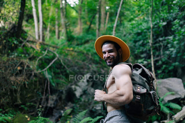 Снизу обтягивающий мужчина-путешественник с рюкзаком и в шляпе, стоящий в скалистом лесу и смотрящий в камеру — стоковое фото