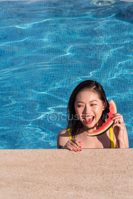 Fröhliche ethnische Frau im Schwimmbad, die an einem sonnigen Sommertag auf einer Wassermelonenscheibe telefoniert und wegschaut — Stockfoto