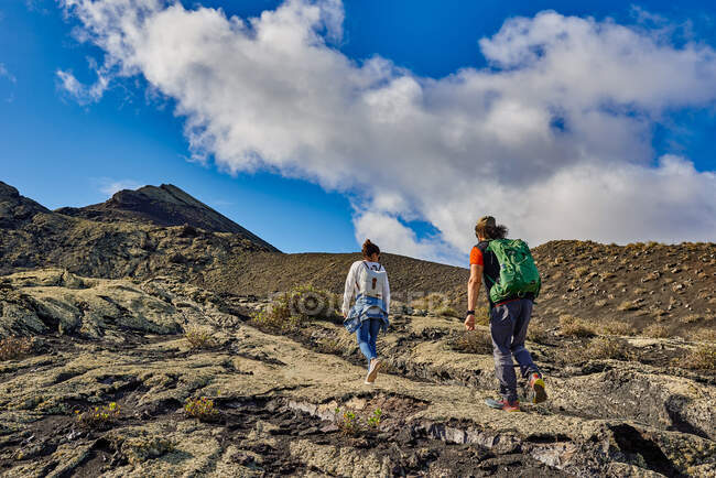 Desde abajo hombre y mujer con mochilas caminando por la ladera de la montaña contra el cielo azul nublado en Fuerteventura, España - foto de stock