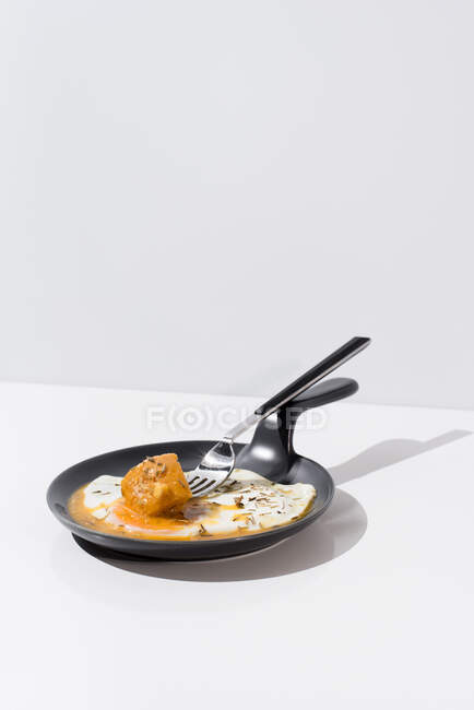 Metal fork with fresh piece of bread dipped in liquid yolk of fried egg served on skillet on white background — Stock Photo