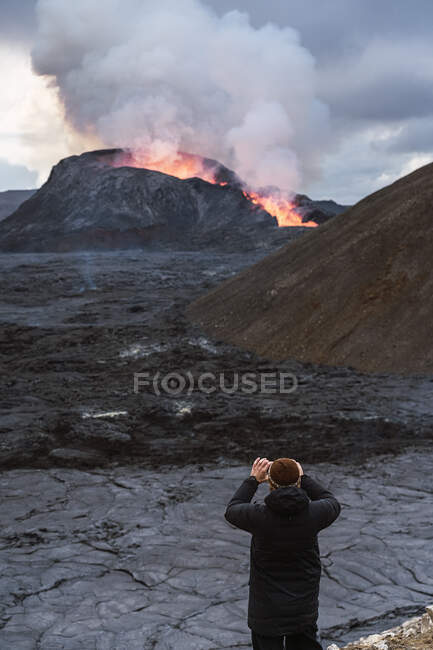 Vista posterior de un viajero irreconocible tomando fotos a Fagradalsfjall con fuego y lava mientras está de pie en el monte en Islandia - foto de stock