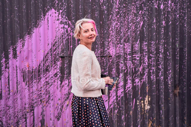 Side view of delighted alternative female with dyed hair and with smartphone standing on background of wall with pink paint in city and looking at camera — Stock Photo