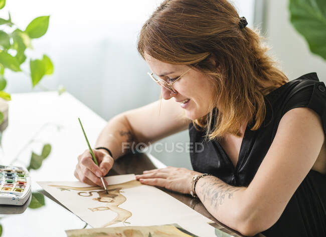 Занятая дизайнерская картина с кистью на бумаге, сидя за столом в креативном офисе и работая над проектом — стоковое фото