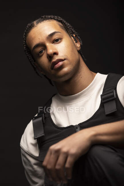 Beau jeune homme ethnique avec des tresses afro habillées en noir et blanc regardant la caméra alors qu'il était assis dans un studio sombre — Photo de stock