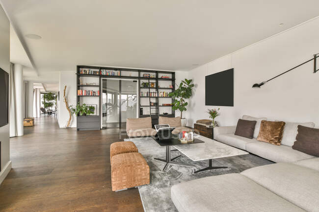 Diseño interior de una elegante sala de estar espaciosa con TV grande colgada en la pared blanca delante de la mesa y sofás decorados con plantas en macetas en un moderno apartamento de lujo - foto de stock