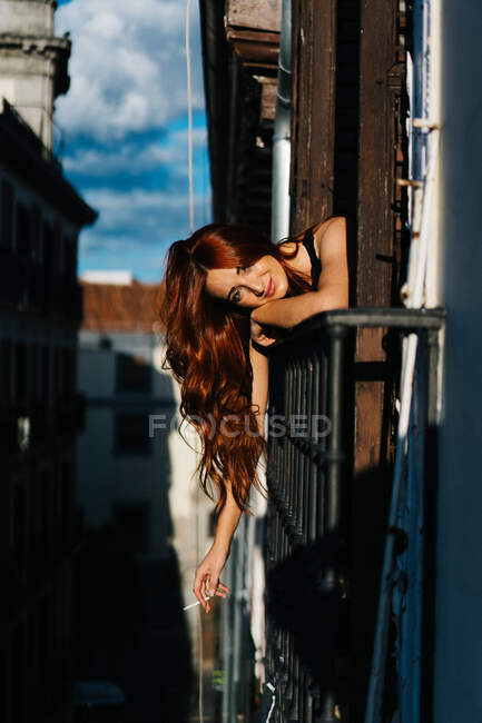 Delighted redhead female with cigarette sticking out of balcony and leaning on metal railing at sunset and looking at camera — Stock Photo