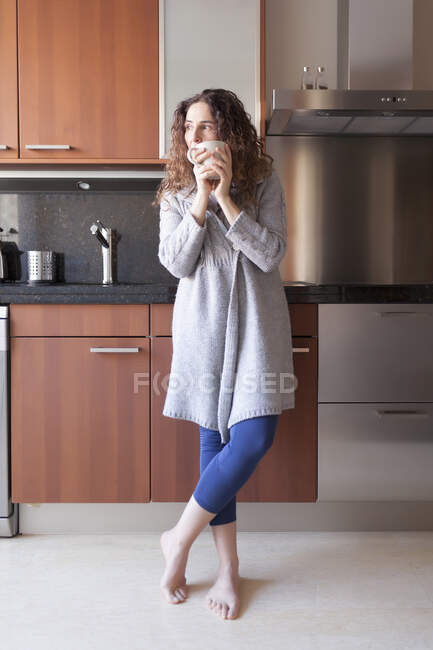 Mujer con el pelo rizado sentada en la cocina tomando una infusión - foto de stock