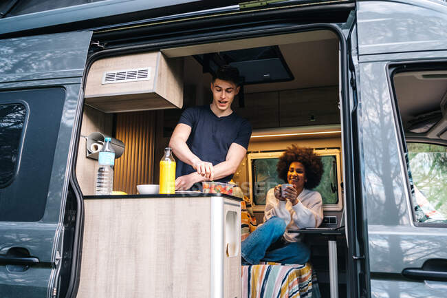 Young guy preparing healthy breakfast while African American girlfriend using gadgets during summer journey together in camper van — Stock Photo