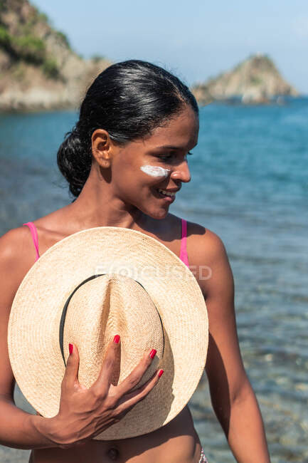 Мечтательная этническая туристка с солнцезащитным кремом на щеках и носом, стоящая со шляпой в руке против моря при солнечном свете — стоковое фото