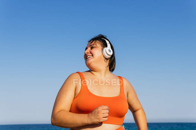 Cheerful ethnic female athlete with curvy body listening to song from headphones under a blue sky — Stock Photo