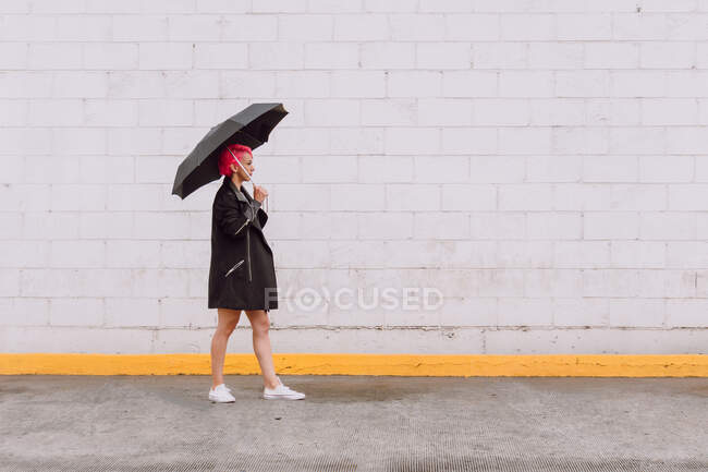 Visão lateral corpo inteiro de jovem fêmea com cabelo rosa na roupa da moda carregando guarda-chuva e andando perto da parede branca na rua — Fotografia de Stock