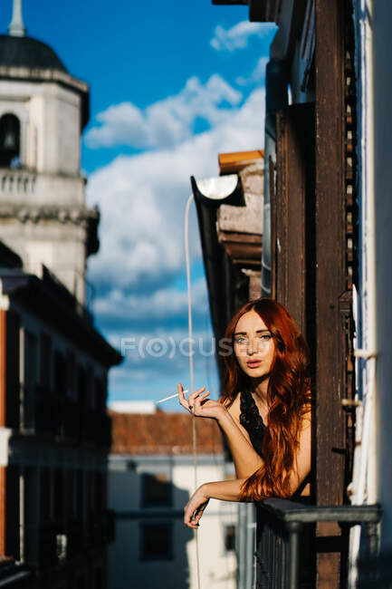 Беззаботная женщина с рыжими волосами, опирающаяся на перила на балконе и курящая сигарету, глядя в камеру в солнечный вечер — стоковое фото