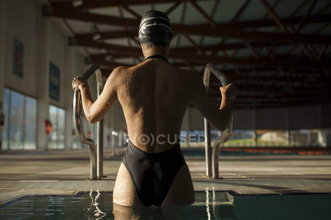 Jovem mulher bonita deixa a piscina interior pelas escadas com maiô preto, vista traseira — Fotografia de Stock