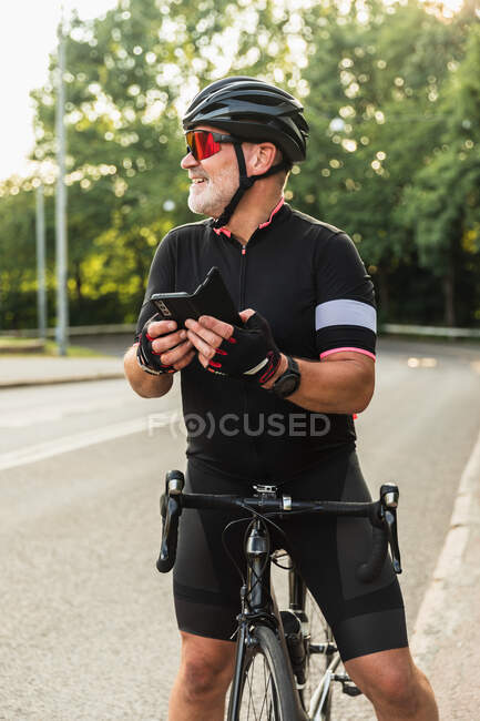 Contenido deportista hispano maduro en casco protector y gafas de texto de mensajería en el teléfono celular mientras está de pie en la carretera urbana con bicicleta - foto de stock