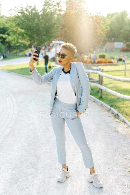 Conteúdo maduro afro-americano feminino em óculos de sol modernos tirando selfie no celular no parque na parte traseira iluminada — Fotografia de Stock