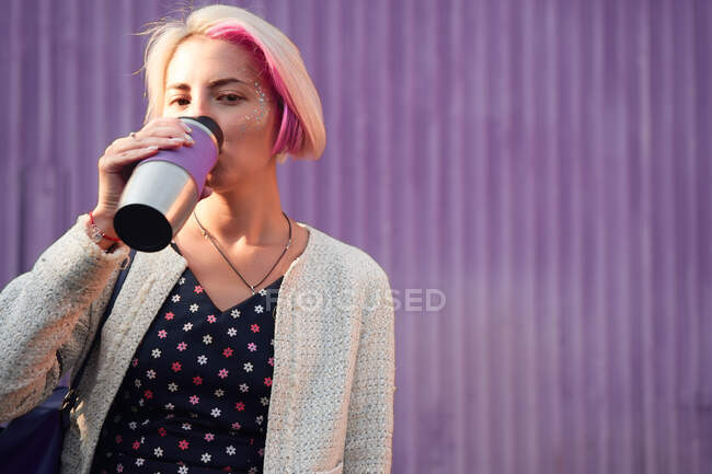 Femme informelle positive avec cheveux courts teints debout avec boisson dans une tasse écologique sur fond de mur violet en ville et regardant la caméra — Photo de stock
