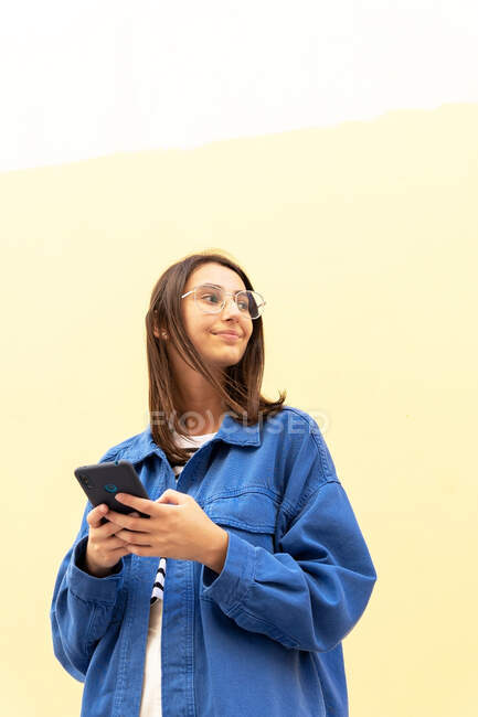 Dal basso della giovane donna in abito alla moda messaggistica sul cellulare sullo sfondo del muro in strada e guardando altrove — Foto stock