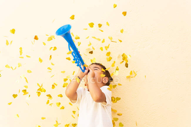 Happy boy in white t shirt standing playing trumpet toy under pile of yellow confetti on light orange wall — Stock Photo