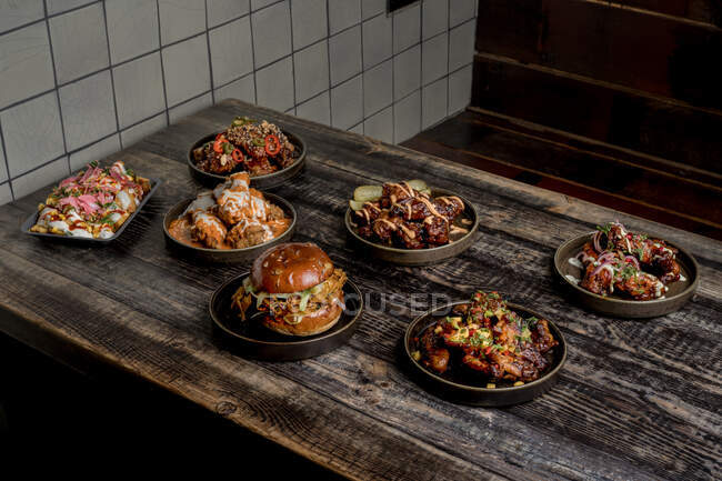 Desde arriba de apetitosas alitas de pollo en salsa BBQ colocadas cerca de hamburguesas y papas fritas con queso sobre mesa de madera en restaurante - foto de stock