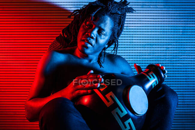 Sognante musicista maschio nero con busto nudo che suona il tamburo africano in studio con luci al neon rosse e blu — Foto stock