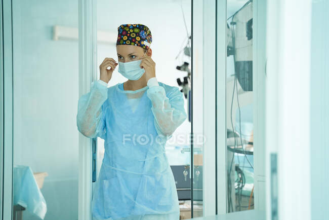 Доросла жінка-лікар у хірургічній формі та орнаментальній медичній шапці, одягнена в одноразову маску, дивлячись вперед у лікарні. — стокове фото
