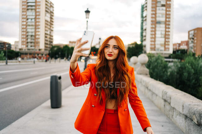 Deliciosa hembra de moda con cabello rojo y traje naranja caminando por la calle en la ciudad y tomando autorretrato en el teléfono inteligente - foto de stock