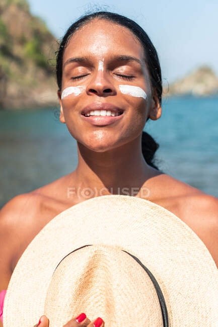Мечтательная этническая туристка с солнцезащитным кремом на щеках и носу, стоящая с закрытыми глазами и шляпой против моря при солнечном свете — стоковое фото