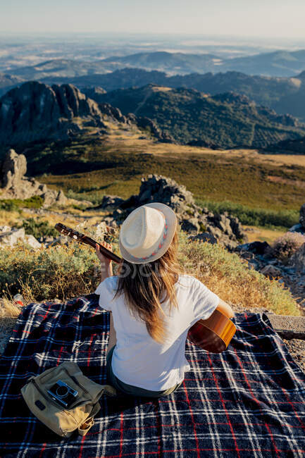Back view of anonymous female traveler in hat sitting on checkered blanket near bag and photo camera and playing acoustic guitar on sunny day in mountains — Stock Photo