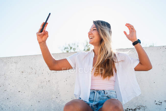 Позитивная жизнерадостная молодая женщина, снимающая себя на мобильный телефон в солнечный день летом в городе — стоковое фото